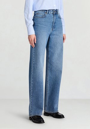 Jeans a gamba larga color azzurro chiaro in denim, con vita alta, passanti per cintura e cuciture a contrasto. Indossati con una camicia a righe blu e scarpe nere.