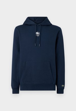 Sweat à capuche bleu marine avec poche kangourou, cordons, et logo blanc Lacoste avec emblème crocodile sur la poitrine et le poignet.