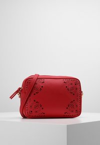 Červená obdélníková crossbody taška s květinovými laserově vyřezávanými vzory a nastavitelním popruhem, vystavená na bílém podstavci na jednobarevném pozadí.