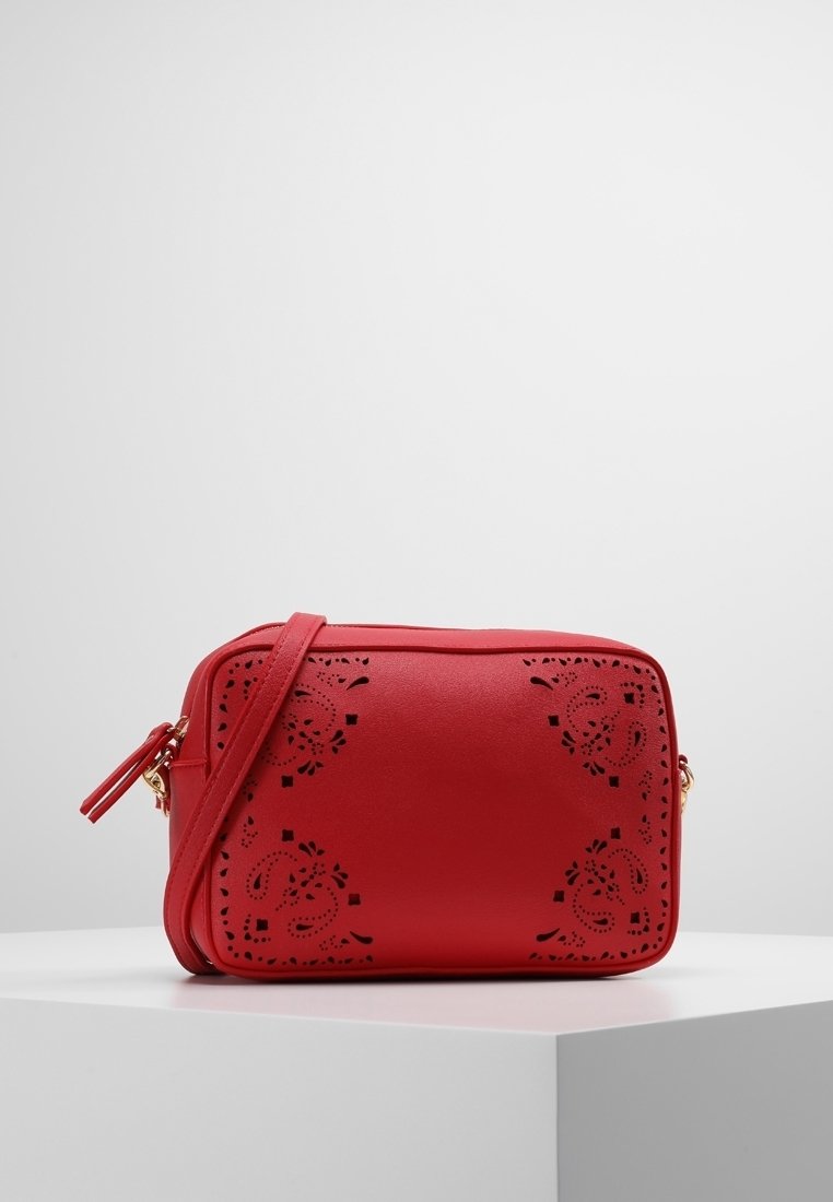 Červená obdélníková crossbody taška s květinovými laserově vyřezávanými vzory a nastavitelním popruhem, vystavená na bílém podstavci na jednobarevném pozadí.