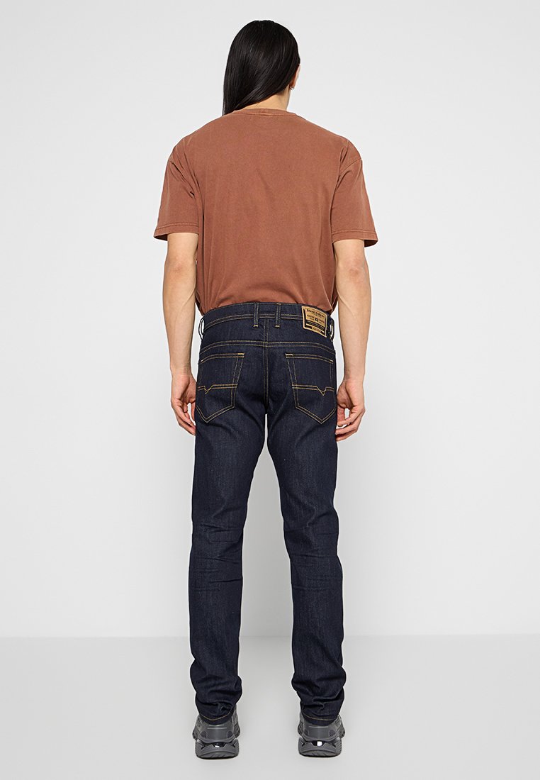 Un adulte avec de longs cheveux bruns portant un t-shirt marron, un jean bleu foncé et des baskets grises se tenant face à un mur gris uni.