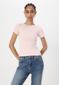 T-shirt ajustée rose clair en tissu côtelé avec des manches courtes. Présente un petit logo brodé sur la poitrine. Associée à un jean en denim bleu.