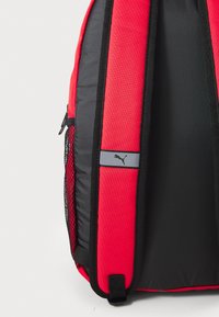 Rucsac roșu și negru, fabricat dintr-un material durabil, având buzunare laterale din plasă, bretele ajustabile și un accent cu logo pe față.