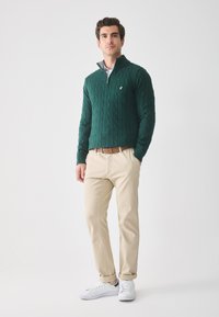 Groene kabelgebreide trui met een kwart-rits kraag, gecombineerd met beige broeken en witte sneakers. Een bruine riem accentueert de outfit.