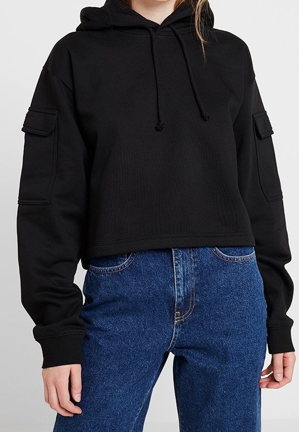 Femme portant un sweat à capuche court noir avec poches avant sur les manches et un jean bleu taille haute sur un fond uni.