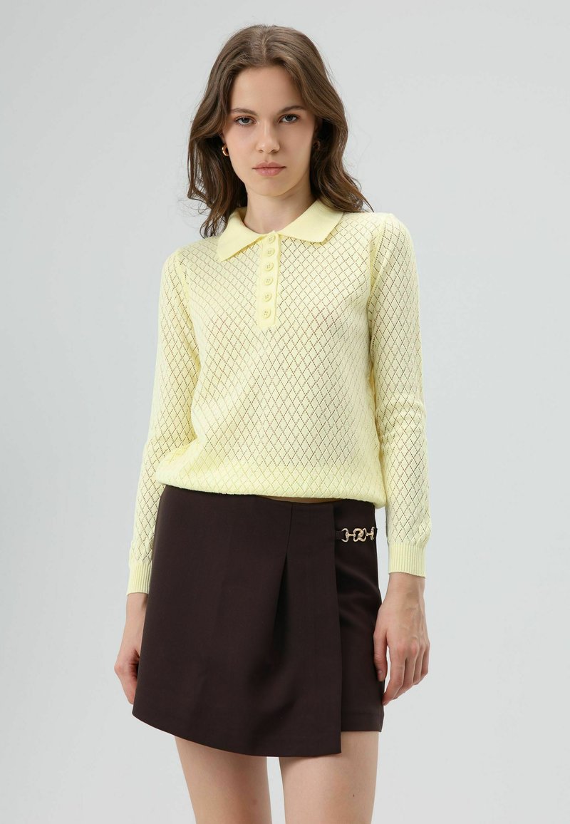 Lichtgeel gebreide trui met een ruitpatroon, kraag en knoopsluiting, gecombineerd met een bruine rok met een gouden kettingdetail.