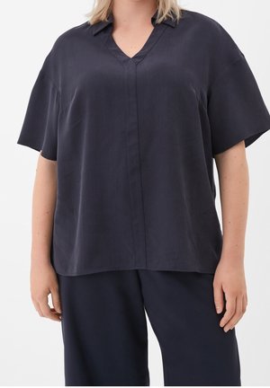 Personne portant un chemisier noir à manches courtes et col en V, ample, associé à un pantalon noir assorti, debout devant un fond blanc.