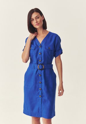 Žena v modrých košeľových šatách po kolená s krátkymi rukávmi a opaskom s okrúhlou prackou, stojaca na jednoduchom pozadí.