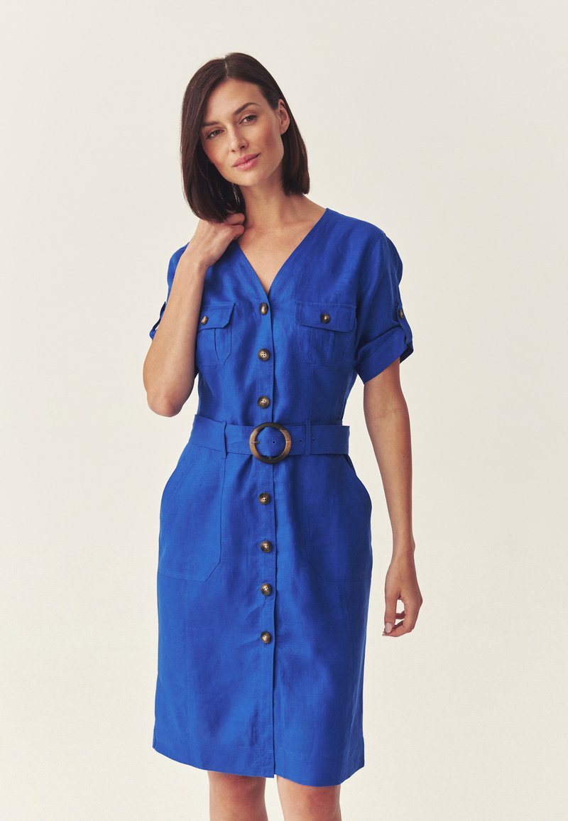 Femme portant une robe bleue boutonnée jusqu'aux genoux, à manches courtes et ceinturée avec une boucle ronde, debout devant un fond uni.