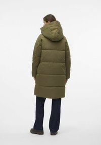 Abrigo acolchado verde con capucha, que presenta un diseño de acolchado y mangas largas. Combinado con pantalones de mezclilla oscuros y calzado grueso.
