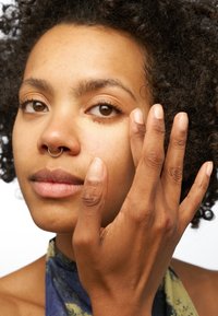 Gros plan d'un visage à la peau brune, avec des cheveux bouclés et un anneau septum doré. La main applique un produit près de l'œil, montrant la texture naturelle.