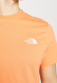 Pomarańczowy bawełniany T-shirt z krótkimi rękawami, z małym białym logo "The North Face" na lewej części klatki piersiowej. Gładka faktura.