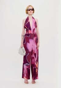 Jumpsuit con collo ad anello di colore viola scuro con motivi floreali in rosa e arancio. La modella tiene in mano una pochette argentata texture e indossa occhiali da sole.