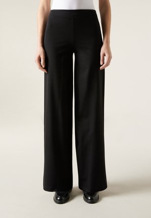 Personne portant un pantalon noir à jambes larges, un haut blanc et des chaussures noires, debout devant un fond clair uni.