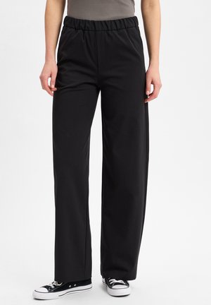 Persoon in een zwart, hoge taille, wijde broek met elastische tailleband en zwart-witte sneakers, staand tegen een witte achtergrond.