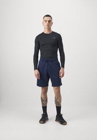 Fekete hosszú ujjú felső és sötétkék sort összeállítás, amely illeszkedő fazonú, rugalmas derékpánttal rendelkezik, és fekete sportcipővel van kiegészítve, gumitalppal.