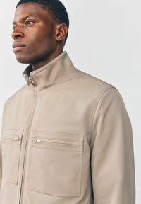 Beige jakke med høj krave, lavet af glat stof, med to brystlommer med lynlåse og synlige syninger.