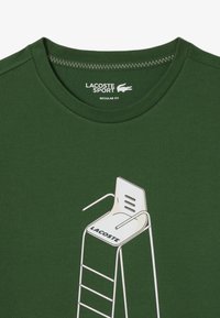 Vihreä puuvillapaita, jossa on valkoinen eloonjäämispenkki graafinen kuvasto ja "LACOSTE" -teksti, suunniteltu tavallisessa istuvuustyylissä.