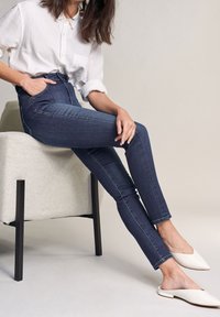Jeans de mezclilla de tiro alto en azul oscuro con corte ajustado, combinados con una camisa blanca de botones y mule beige, sentada en una silla de color claro.