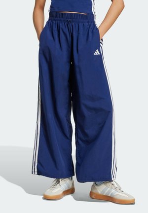 Osoba ubrana w luźne granatowe spodnie dresowe Adidas z białymi paskami po bokach oraz białe trampki z gumowymi podeszwami, stojąca z rękami w kieszeniach.