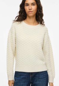 Pull beige clair texturé avec un motif en nid d'abeille, col rond et manches longues, associé à un jean bleu. Matière en maille douce.
