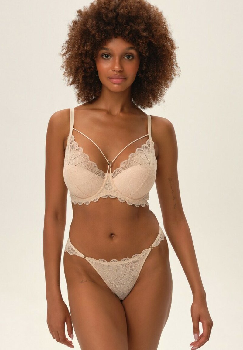 Beżowy komplet biustonosza i stringów z koronką, ozdobiony kwiatowymi wzorami, falbankami i regulowanymi ramiączkami. Biustonosz ma centralny dekoracyjny akcent.