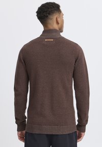 Brauner Strickpullover mit hohem Kragen, strukturiertem Muster, gerippten Bündchen und Saum. Verfügt über ein kleines Markenzeichen auf der Rückseite.