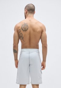 Szürke úszónadrág sima textúrával, oldalzsebekkel és kontrasztos varrással. Egy személy hátulnézetből viseli.