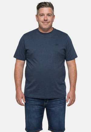 Homme portant un t-shirt à manches courtes bleu marine et un short en denim foncé, se tenant devant un fond gris clair uni, souriant doucement.