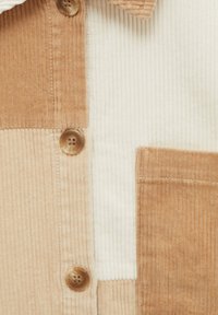 Gros plan d'une chemise en velours côtelé avec des blocs de couleurs beige, sable et crème, avec des boutons marron et une poche poitrine sur le côté droit.