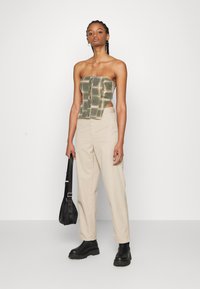 Crop top sans manches à motif vert avec des boutons, associé à un pantalon taille haute beige. Le modèle tient un sac noir et porte des bottes noires imposantes.