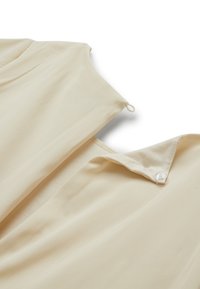 Detailní záběr na béžovou látku s límečkem, který má průhledný knoflík a malý detail smyčky na bílém pozadí.