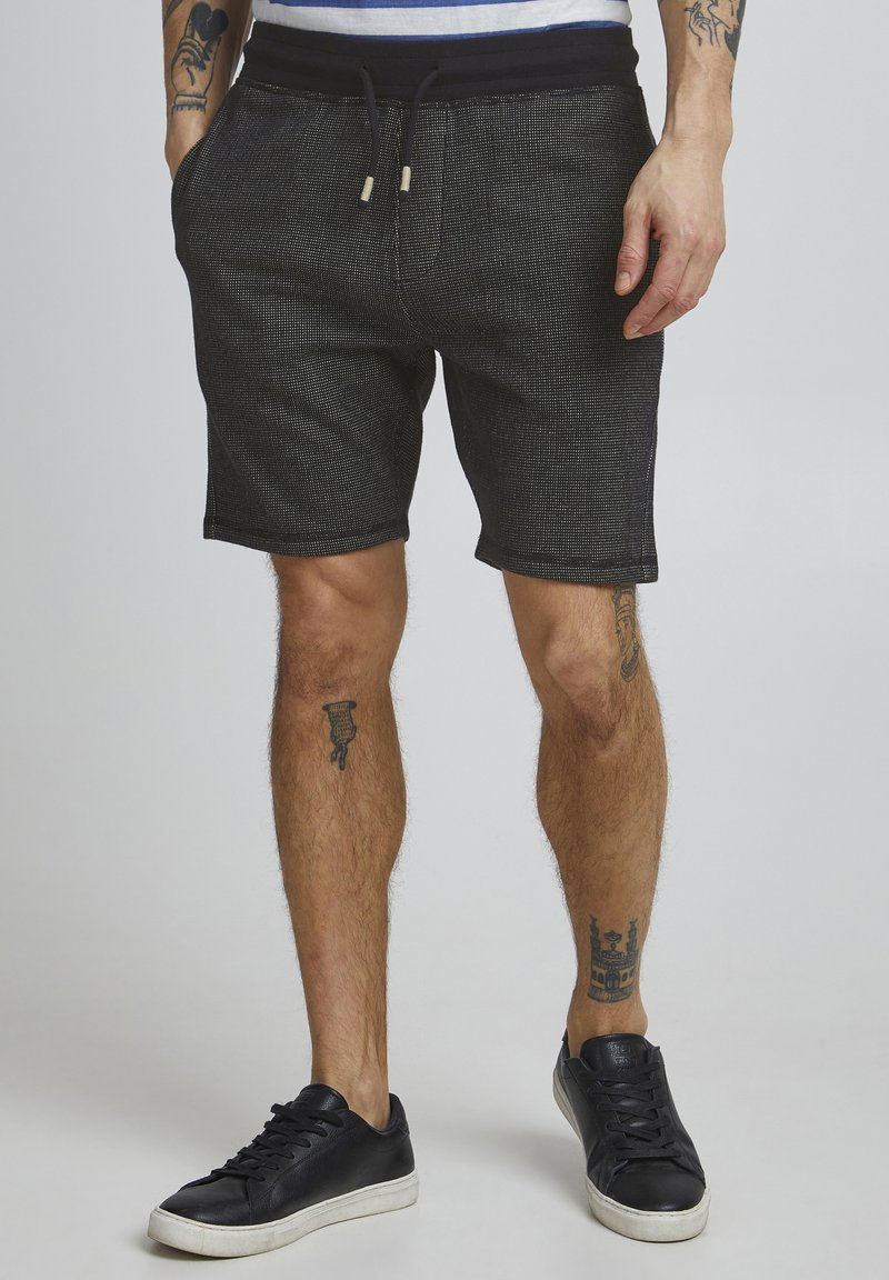 Shorts noirs texturés avec une taille élastique à cordon de serrage et des poches, associés à des baskets en cuir noires avec des semelles blanches.