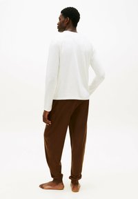 Uomo in piedi scalzo di spalle, indossa una camicia bianca a maniche lunghe e pantaloni marroni larghi su sfondo bianco.