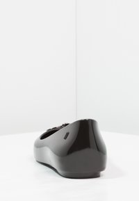 Zapato plano negro con superficie lisa y brillante. Tiene una punta redondeada y un leve tacón. El detalle lateral incluye una textura de línea vertical.