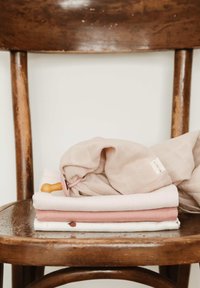 Pehmeatest, tekstuuriga puuvillastes musliinrätikutest virn beeži ja roosa toonides, alumisel riidetükil väike mustriline detail, mis puhkevad puidust toolil.