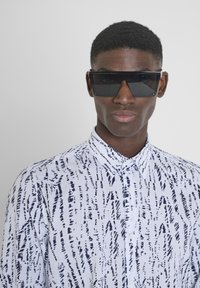 Occhiali da sole neri oversize e una camicia bianca con bottoni con un motivo astratto blu navy. Capelli corti e texturizzati visibili.