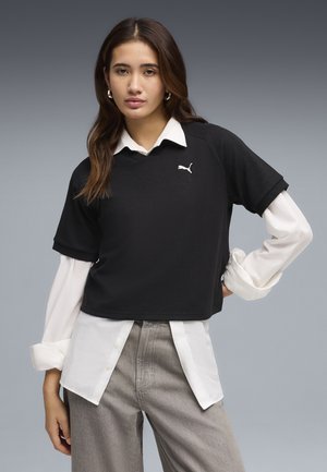 Jeune femme aux longs cheveux bruns portant une chemise blanche à plusieurs couches et un pull noir à manches courtes avec le logo Puma, debout devant un fond gris.