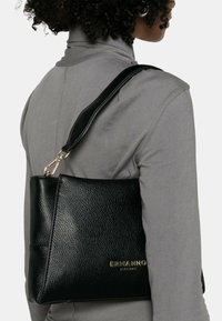 Sac à main en cuir noir avec une finition texturée, arborant un logo doré à l'avant et une bandoulière détachable et réglable.