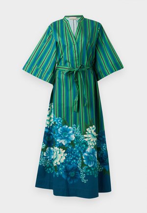 Vrouw draagt een groen-blauwe gestreepte jurk met bloemenprint aan de zoom, houdt een kleine zwarte en beige geweven handtas vast.