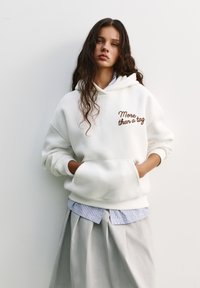 Felpa bianca con cappuccio e testo ricamato marrone "More than a tag", dotata di tasca frontale e indossata sopra una camicia a righe blu e bianca.
