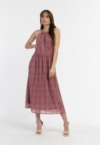 Punainen kuviollinen halterneck-mekko, jossa on kerrottu vyötärö, virtaava kangas ja polvipituinen helma. Yhdistettynä kirkkaisiin korkokenkiin ja renkaisiin korvakoruihin.