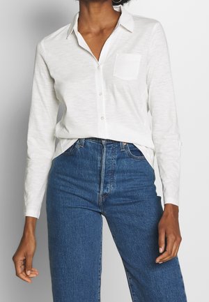 Personne portant une chemise blanche à manches longues avec une poche poitrine et un jean bleu taille haute sur un fond uni.