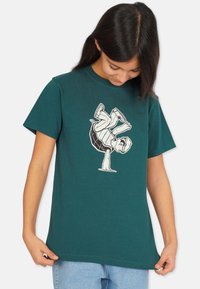 Teal puuvillapaita, jossa on valkoinen graafinen kuvitus kilpikonnasta, joka tasapainoilee kuorensa päällä, lyhyet hihat ja rento malli.