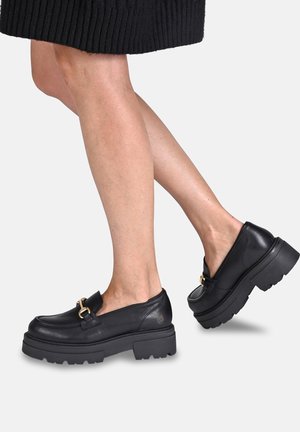 Nogi w czarnych masywnych loafersach na platformie z złotymi klamrami i czarnej prążkowanej spódnicy na jasnym tle.