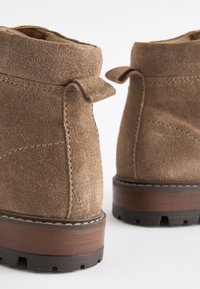 Hnedé semišové členkové topánky s mäkkou textúrou, pevnou gumovou podrážkou a malými textilnými slučkami na vrchu pre jednoduché obúvanie.