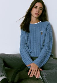 Junge Frau mit langen dunklen Haaren, die einen blauen Zopfmuster-Pullover und graue Jeans trägt, sitzt im Schneidersitz an einer weißen Wand.