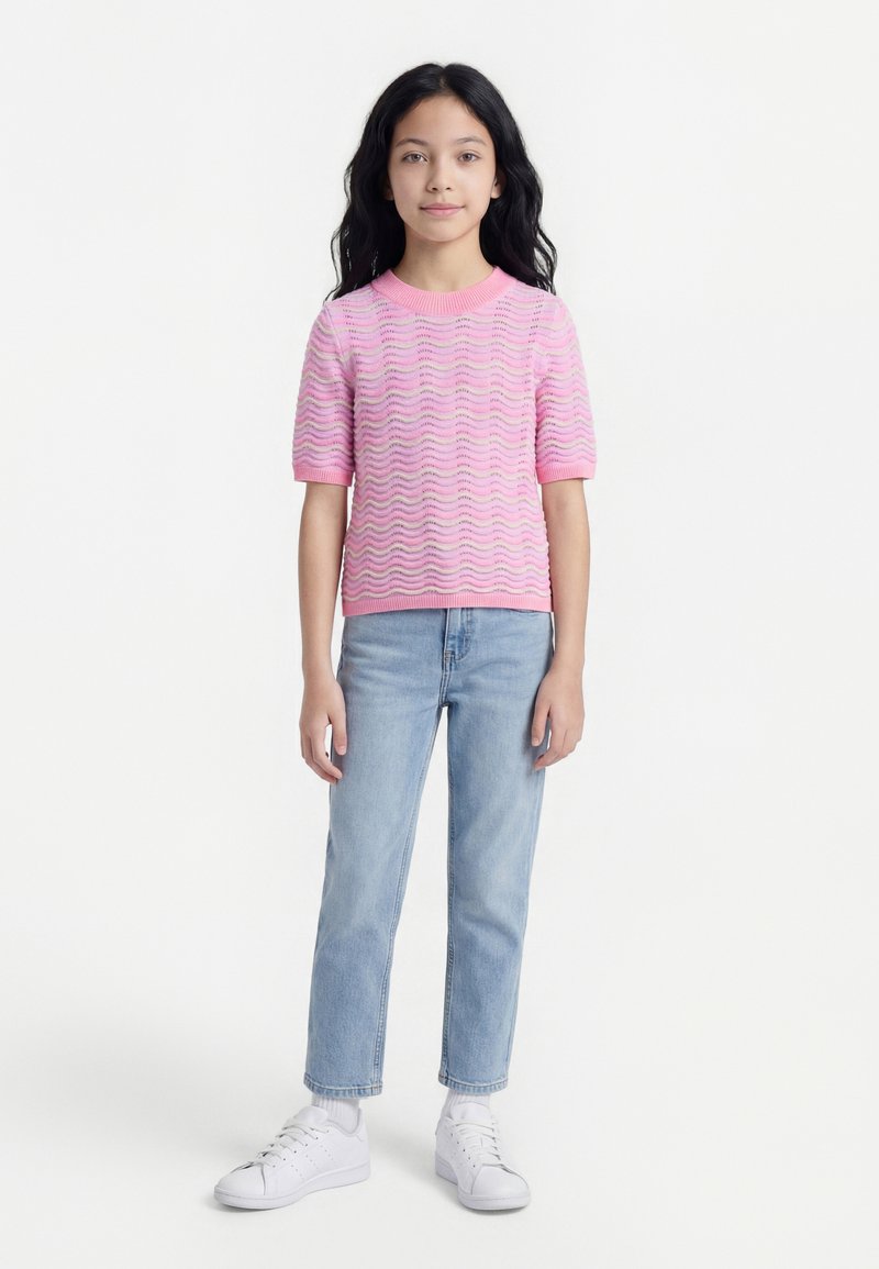 Κοπέλα με μακριά μαύρα μαλλιά, που φοράει ροζ πουλόβερ με κοντά μανίκια, ανοιχτό μπλε τζιν, λευκές κάλτσες και λευκά αθλητικά, στέκεται μπροστά σε λευκό φόντο.