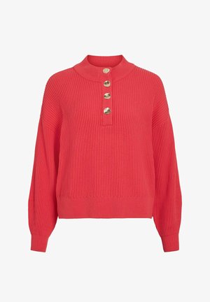 Koraļļu krāsas džemperis ar ribotu audumu, augstu apkakli un četriem zelta toņa pogām pie apkakles. Raksturojams ar vaļīgām, garām piedurknēm un brīvu piegriezumu.