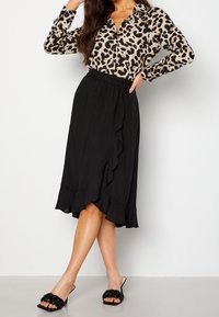 Vrouw in een lange mouw blouse met luipaardprint, gestopt in een zwarte midi rok met ruffles, met zwarte gevlochten open teen hakken.